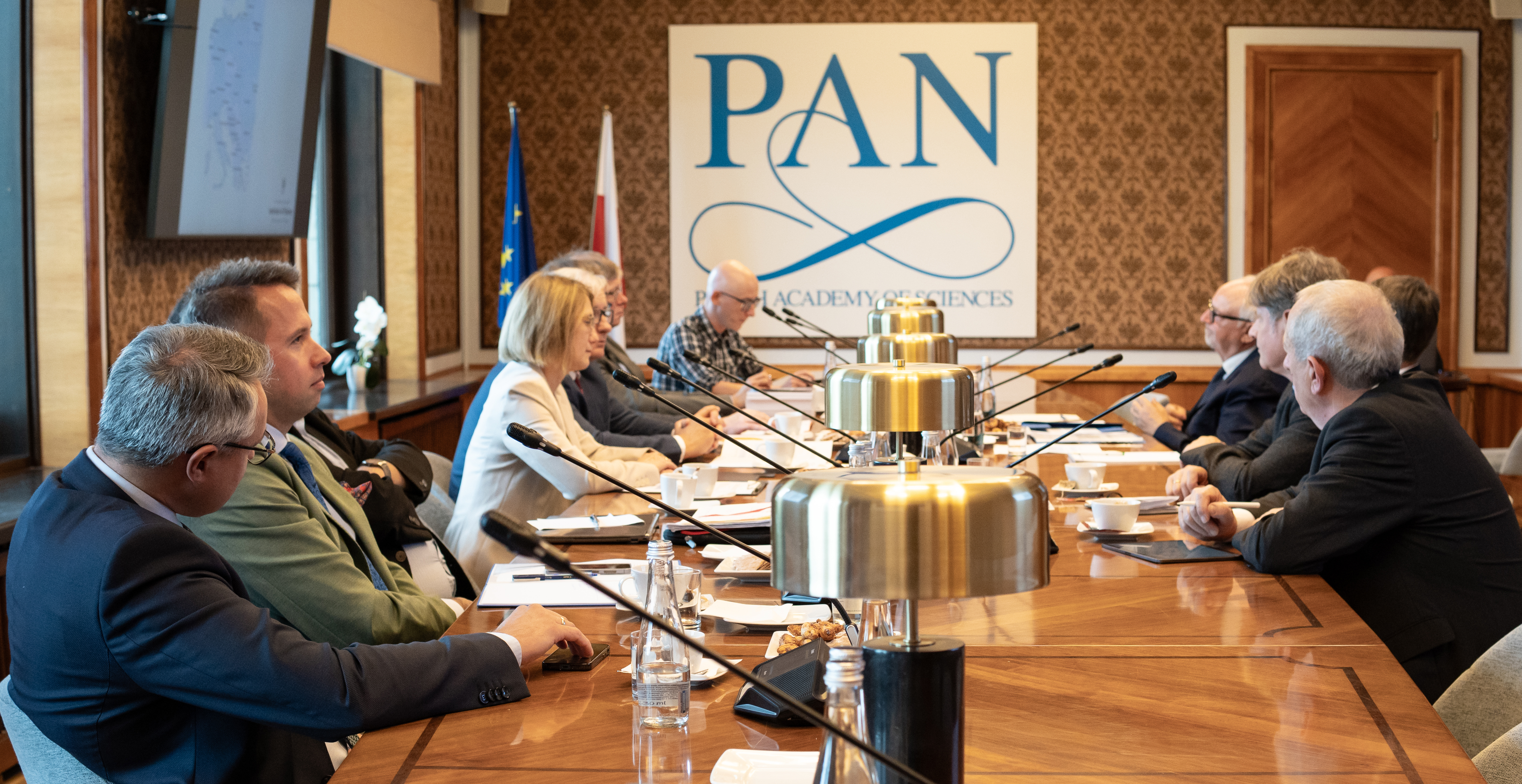 Mehrere Menschen sitzen an einem großen Tisch vor Mikrofonen und sprechen miteinander. Im Hintergrund sieht man einen Aufsteller mit der Aufschrift PAN.