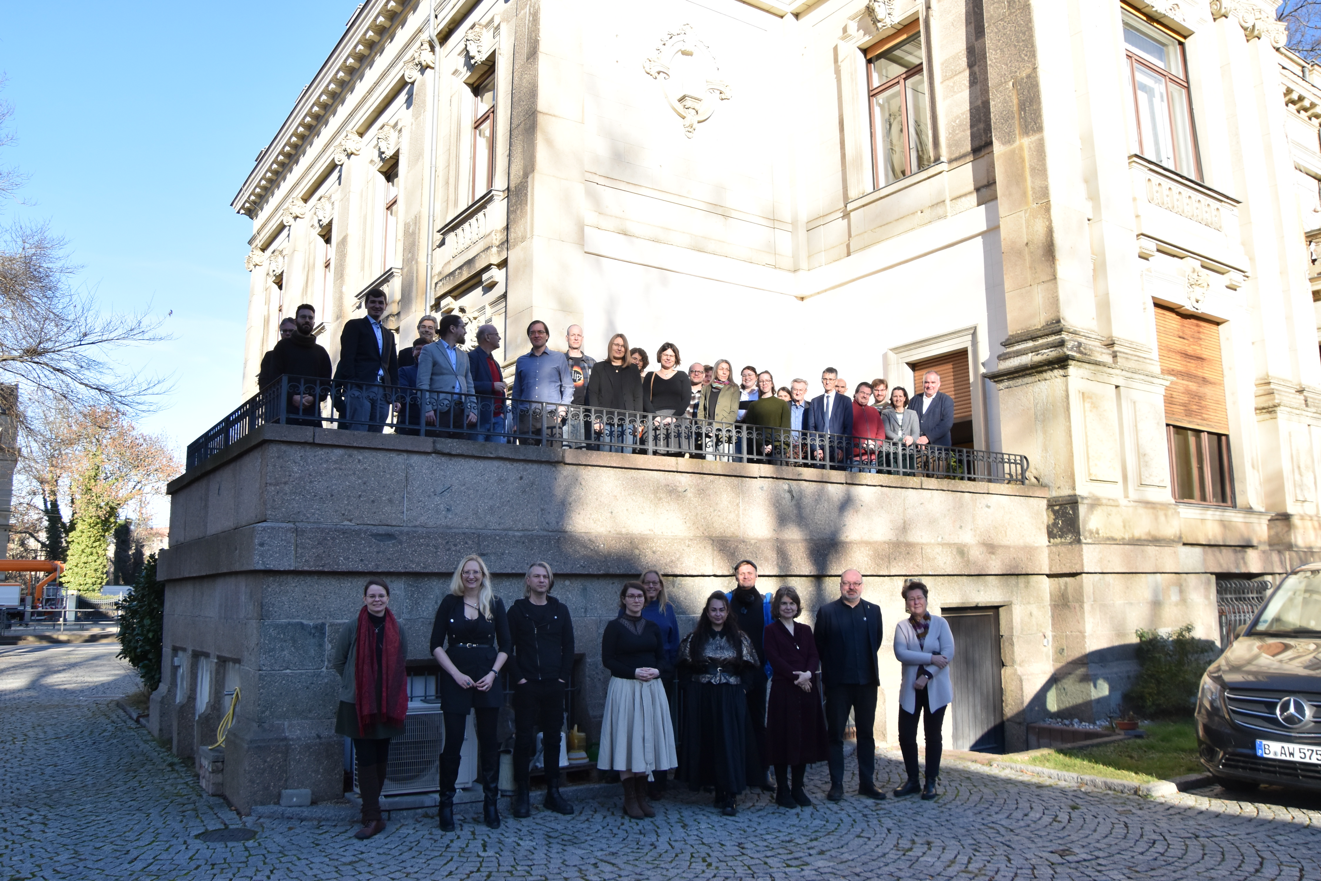 Eine größere Gruppe von Personen steht bei sonnigem Wetter auf und vor der Terasse der Sächsischen Akademie der Wissenschaften.