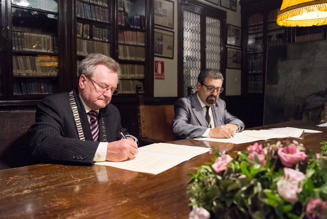 Zwei Männer sitzen an einem Holztisch und unterzeichnen jeweils ein Papier. Im Hintergrund sieht man Bücherregale.