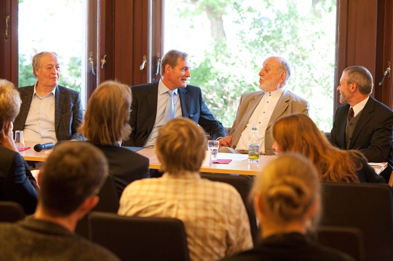 Mehrere männliche Personen sprechen und lachen an einem Tisch sitzend. Im Vordergrund sieht man Publikum sitzen.