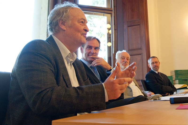 Eine männliche Person gestikuliert am einem Tisch sitzend, hinter ihm sitzen drei weitere Männer und hören zu.