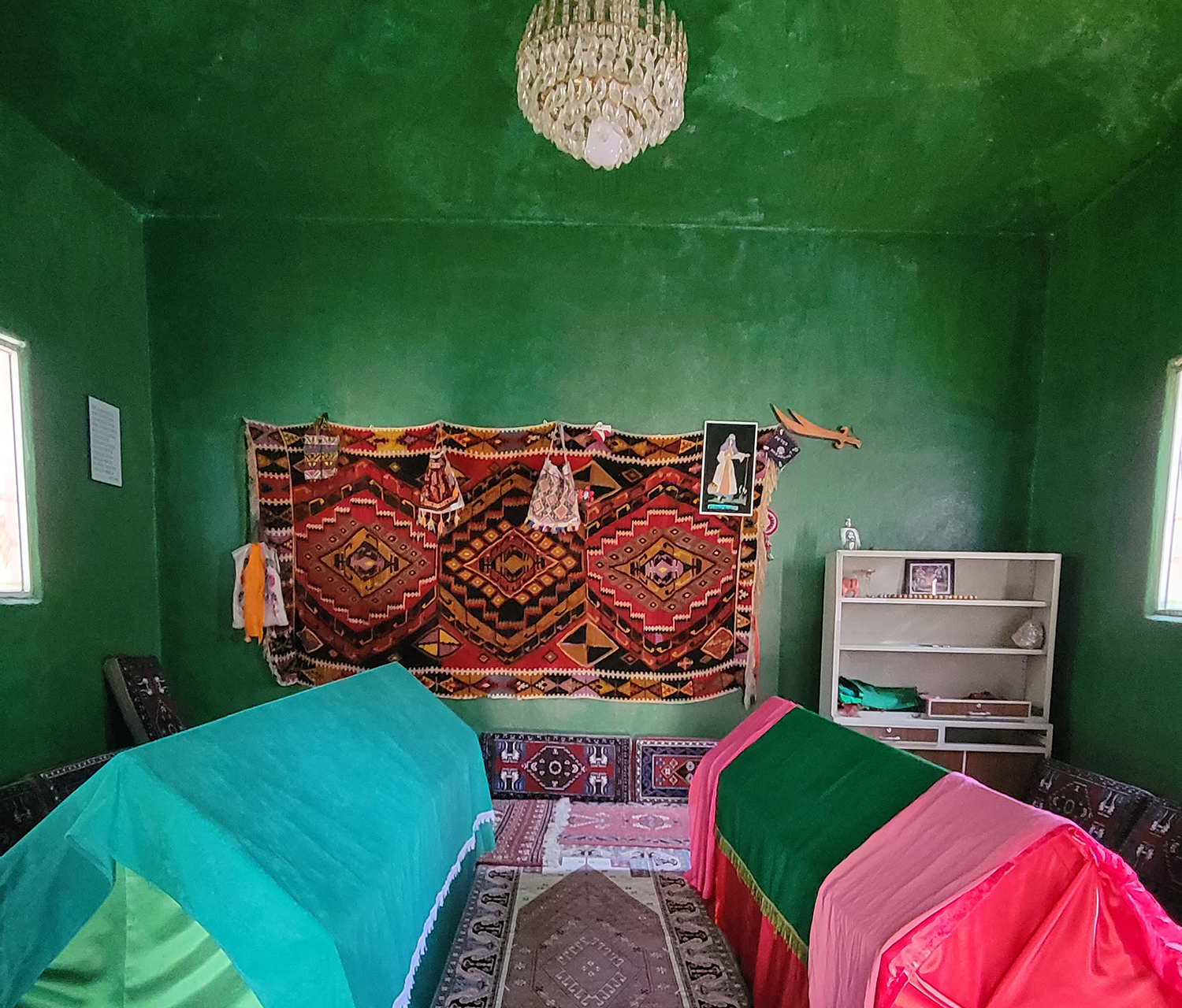 The interior of a mausoleum, with green walls, carpets on the floor and a tapestry on the wall; two coffins are draped in colourful cloths.