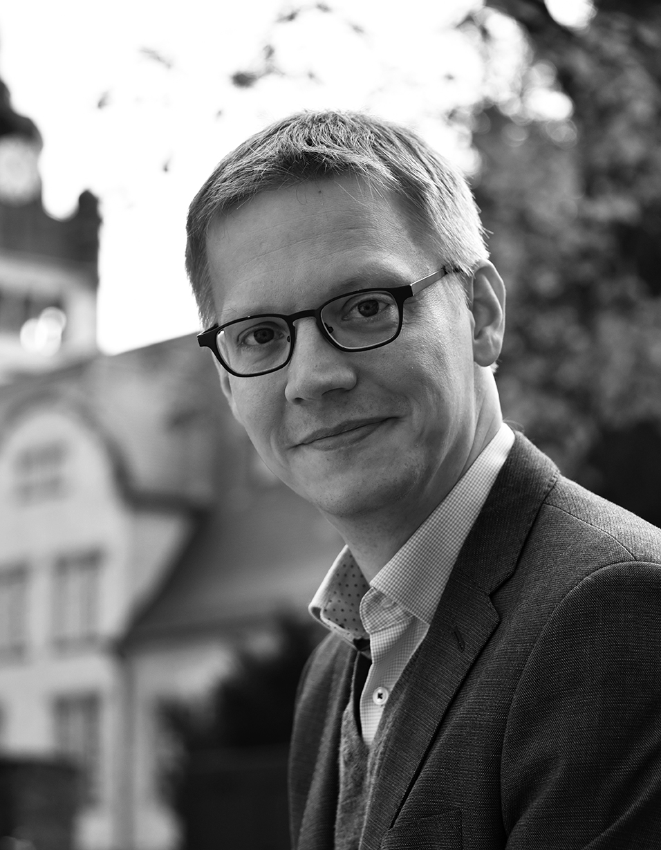 Black-and-white portrait photograph of a smiling man wearing glasses and a suit, standing in front of a historic building
