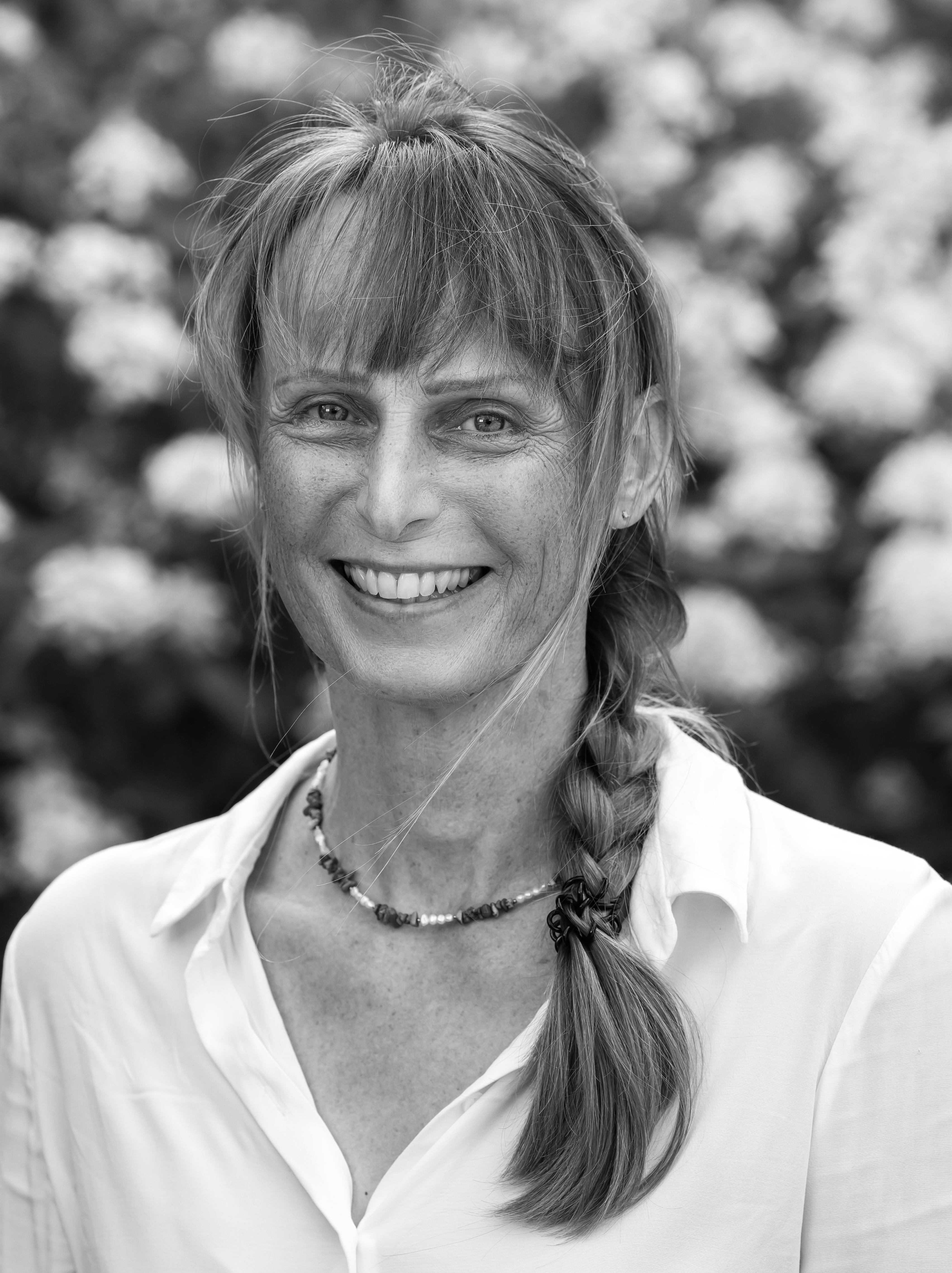 A black-and-white portrait of a smiling woman wearing a white blouse and a necklace, set against a natural backdrop